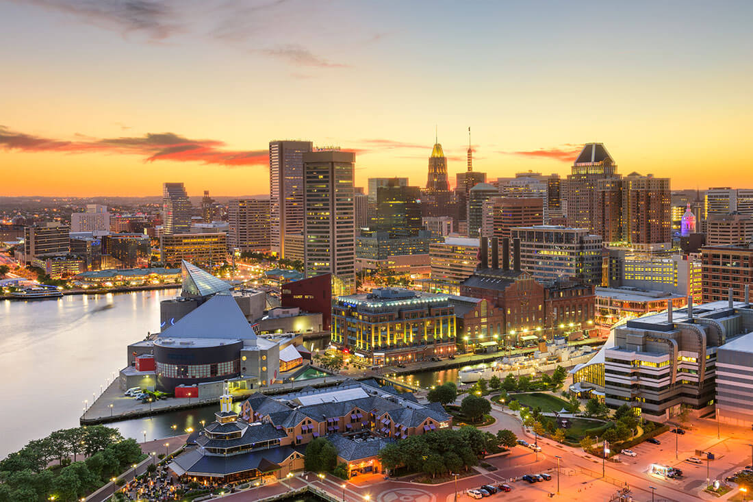 A view of Baltimore's Inner Harbor, framed by a sunset and skyline filled with tall office buildings, cultural attractions and retail establishments.