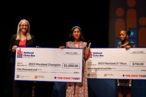 A close up of the middle school students who earned first and second place in the 2023 National Civics Bee for Maryland standing on a stage and both holding a large check for their grand prize earnings, with Whitney Harmel, Executive Director of the Maryland Chamber Foundation, standing with the first place winner.