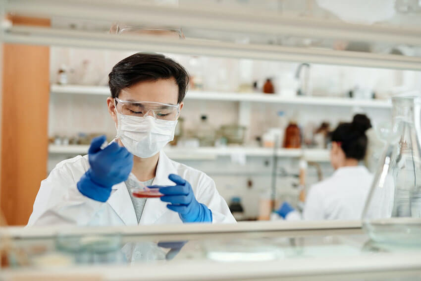 An individual wearing safety glasses, a white mask, a white lab coat and blue gloves uses a test tube and petri dish while a person in the background stocks shelves with medical items.