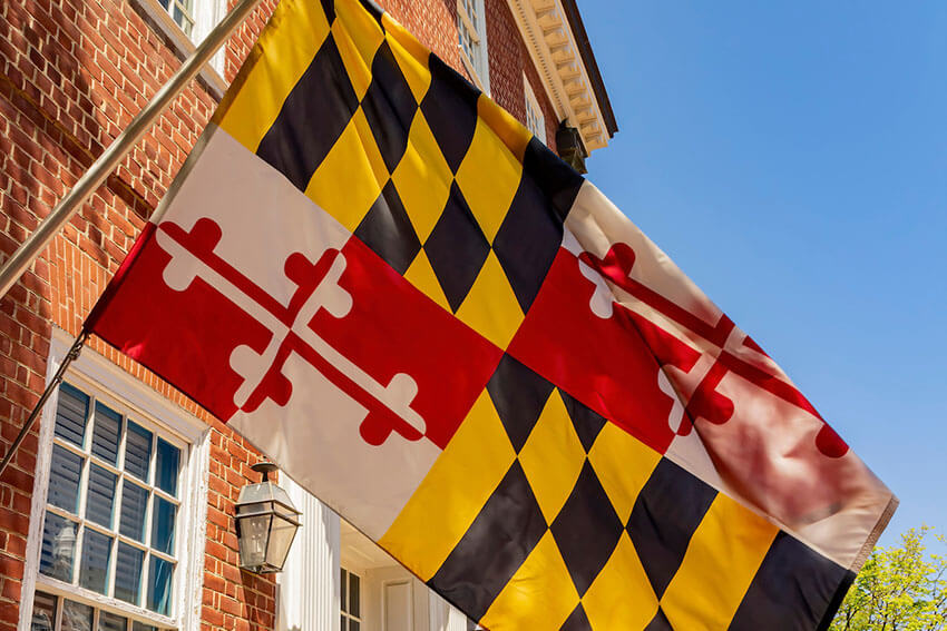 A photo of the Maryland Flag waving in front a bright blue sky with a brick building in the background.