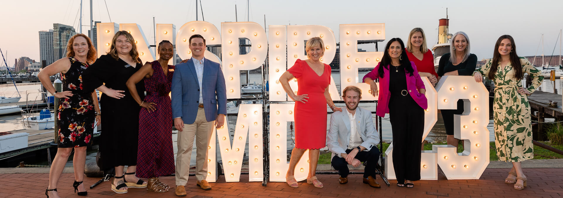 A group of 10 Maryland Chamber of Commerce staff pose in front of of a glowing, 3-D Inspire MD sign, with boats and waterfront from Baltimore's Inner Harbor in the background /
