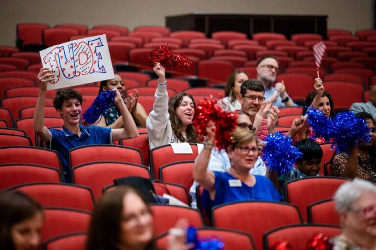 A look at the audience seated in red theater seating and holding up signs, pom-poms and small American flags while cheering on the finalists participating in the 2023 National Civics Bee competition for Maryland.
