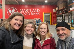 Four individuals pose and smile while standing in the Maryland Food Bank warehouse. A large red wall with a giant Maryland Food Bank logo is in the background, as are pallets and shelves to store food donations.