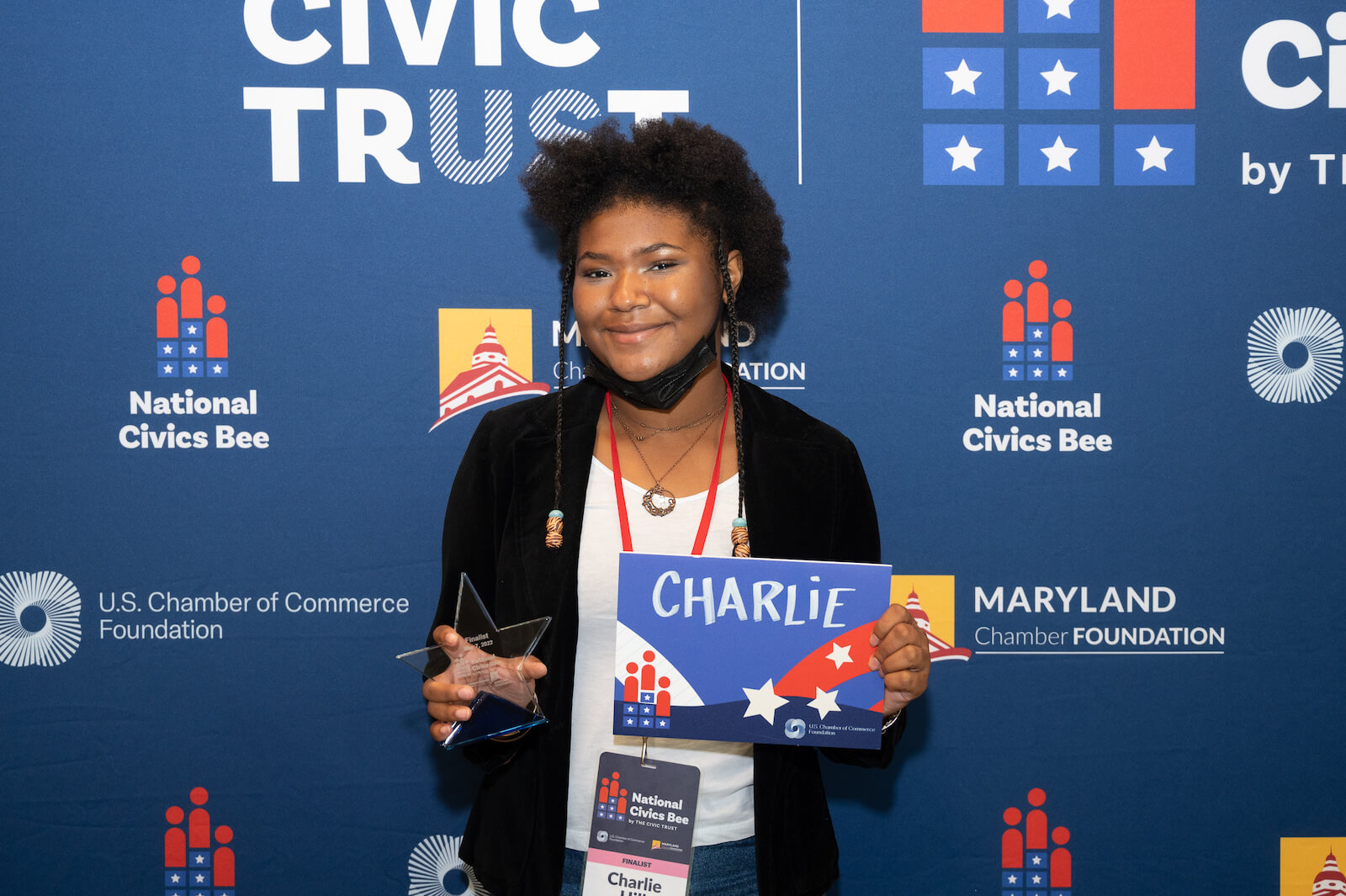 A close up of a middle school student who was a finalist in the 2023 National Civics Bee for Maryland standing in front of a backdrop with the National Civics Bee, U.S. Chamber Foundation, Civic Trust and Maryland Chamber Foundation logos and holding a sign with her name on it.
