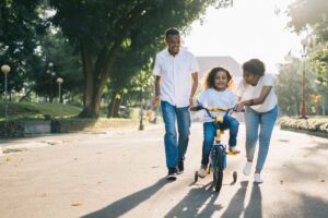 Two adults guide a child who is learning to ride a bike with training wheels on a paved road lined by trees.