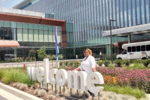 Teacher Extern Alethea Smith standing in front of a 3-D welcome sign at a Kaiser Permanente medical facility, with flowers and landscaping in the background.