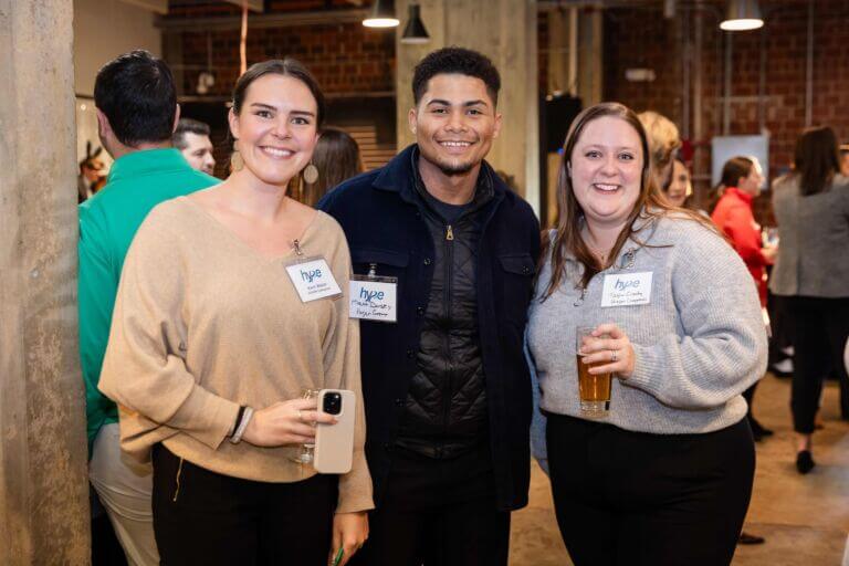 Three individuals smile and pose for a photo while attending the Maryland Chamber's HYPE: Helping Young Professionals-Excel event at Guinness brewery. Several people are shown standing and talking in the background.
