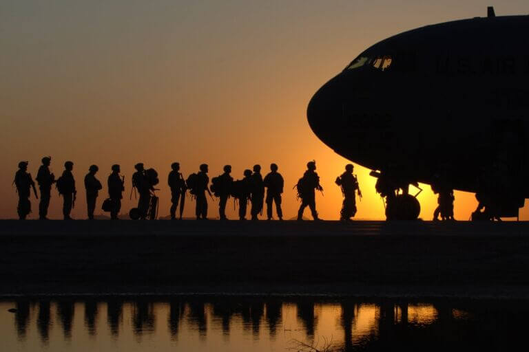 A line of military members stand in front of a plane as they prepare to board, with a setting sun in the background and the nose of a cargo plane showing.