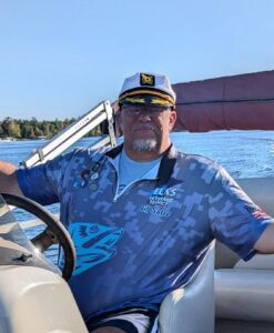 A man driving a pontoon with a captain's hat and a printed shirt for MN Elks Veteran Service.