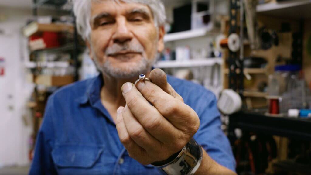 A man with gray hair and blue shirt holds a silver ring with an embedded blue stone.
