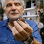 A man with gray hair and blue shirt holds a silver ring with an embedded blue stone.