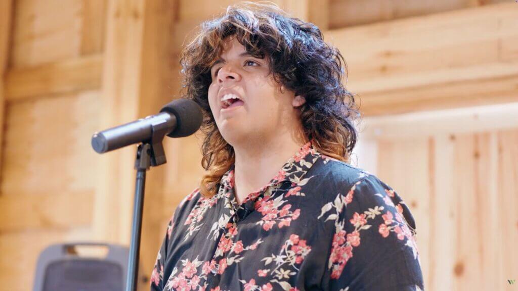 A boy with long black hair and orange highlights sings into a microphone. He is wearing a black shirt with pink and orange flowers on it.