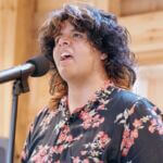 A boy with long black hair and orange highlights sings into a microphone. He is wearing a black shirt with pink and orange flowers on it.