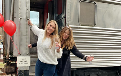 2 woman leaning out of train car at wine tasting