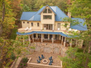 A modern house with a blue metal roof, surrounded by trees, featuring a spacious deck and stone patio with seating.