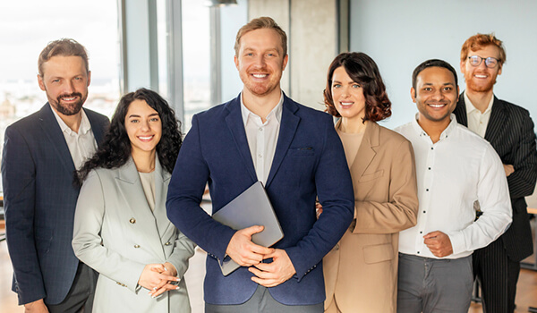 A group of smiling business professionals stand together in an office setting. The man in the center of the group is holding a laptop, while the others stand around him with their arms crossed