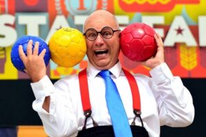 A comic in suspenders holding blue, yellow, & red volley balls in line with his bald head.