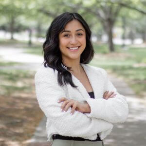 Young woman standing with her arms crossed and smiling in a white top against a blurred outside background