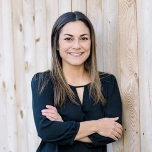 Smiling woman stands with her arms crossed in a black top against a wood grain background.