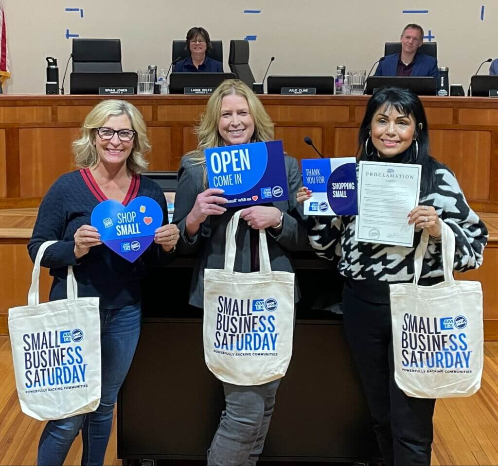 Three women standing next to one another in a court house with signs and bags that say Small Business Saturday