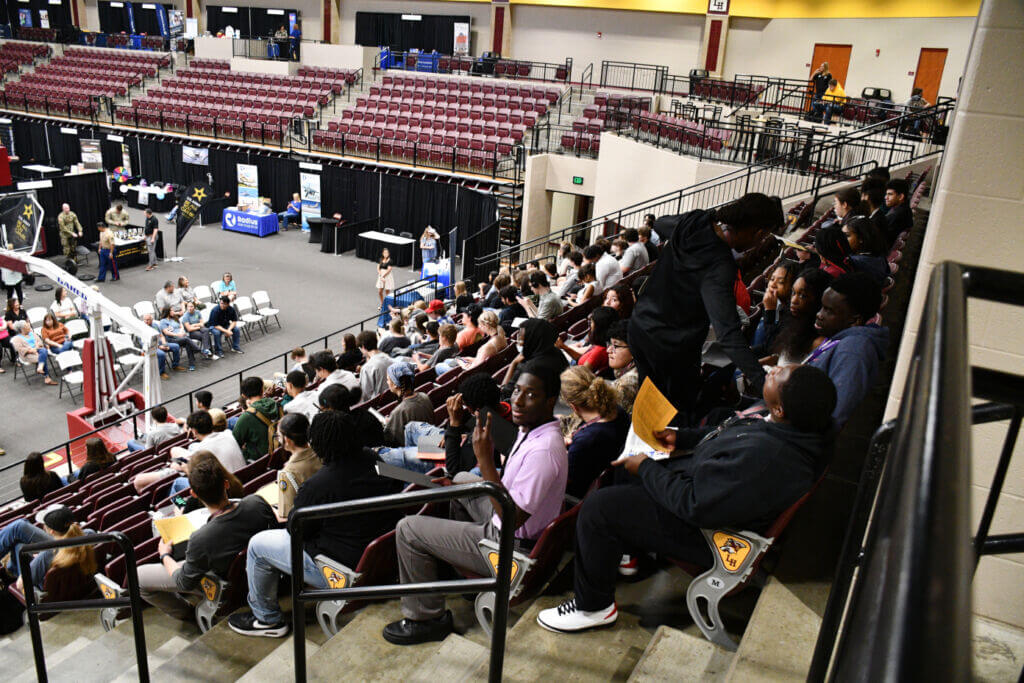 High school students seated in bleachers listen as teachers and industry leaders speak during the opening session of Draft Day 2023.
