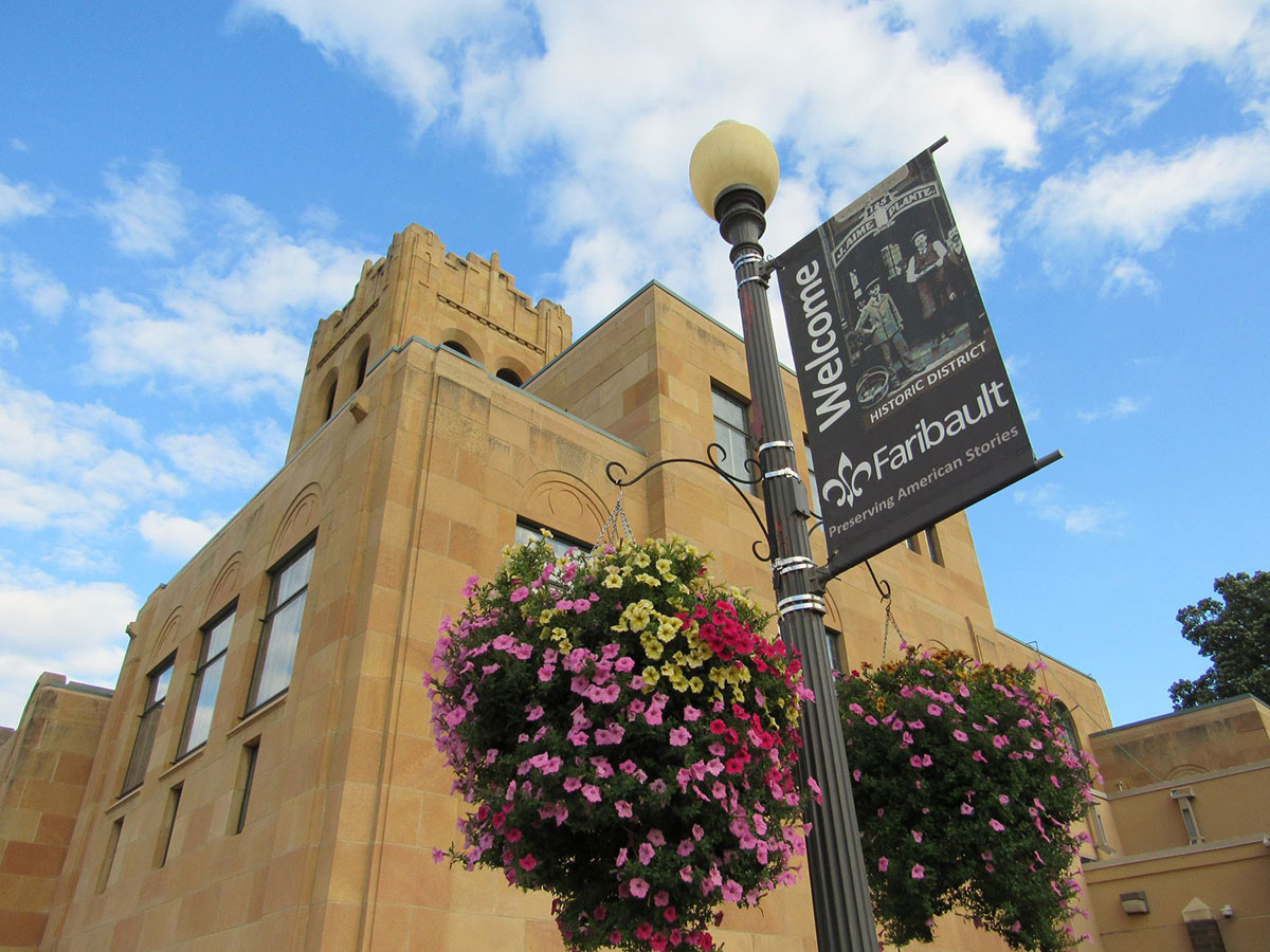Greek inspired library building with banner on pole in front saying welcome to Faribault, surrounded by flower baskets