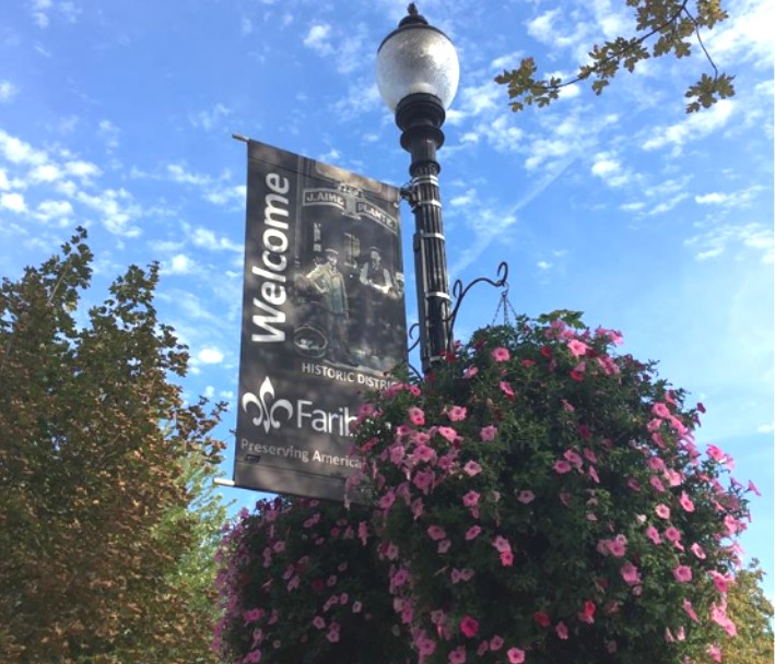 Flower baskets hang from light pole with banner reading welcome to Faribault