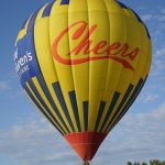 People riding in a hot air balloon that says "Cheers" on the outside of the balloon - Galt Balloon Festival