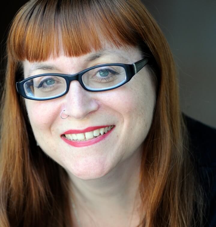 Headshot of Melissa Hillman. A light skinned woman with blue eyes, black oval glasses and wearing red lipstick with a nose ring. She has red hair with bangs. She is smiling with her teeth showing.