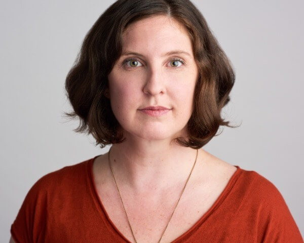 Headshot of Ann Marie wearing an orange shirt and a silver necklace. She is a white woman with chin length short brown hair.