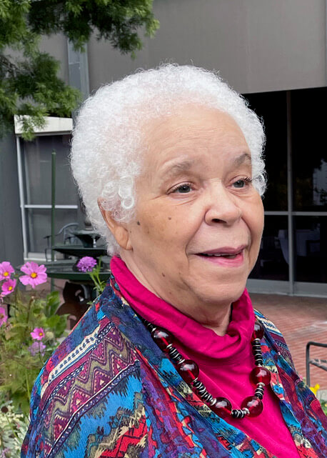 Anne Smith in a colorful sweater smiles, looking off to the right of the camera. She is a Black woman with short white hair.