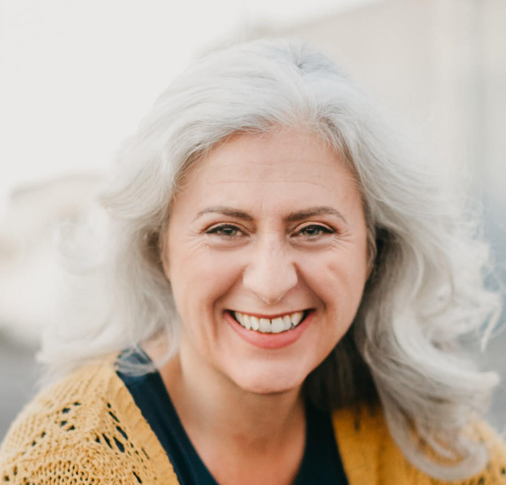Headshot of Debórah Eliezer. She is a white woman with white hair wearing a black shirt with a yellow sweater. She is looking at the camera and smiling with her teeth showing.