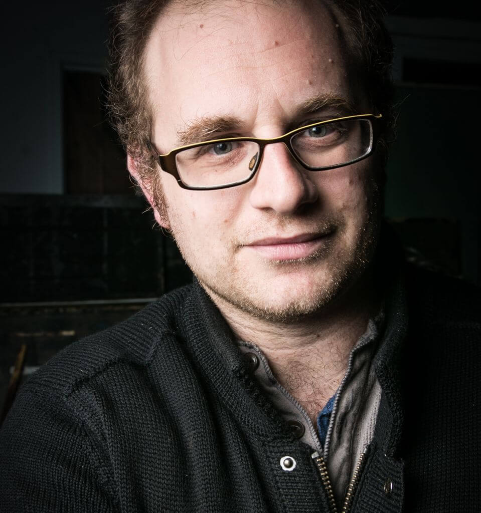 A white partially balding human with glasses looking into the camera. He is wearing a dark grey sweater over a grey buttoned shirt