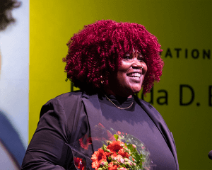 Rolanda D. Bell, a Black woman with red curls, smiles in the spotlight, holding a bouquet of flowers.