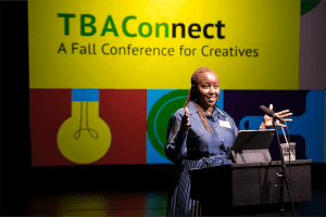 Nataki Garrett, a Black woman in a blue dress, gestures at a podium, with the TBAConnect graphic projected in the background.