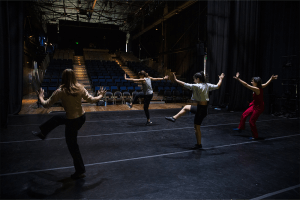 Four people in a line practicing a dance move on stage.