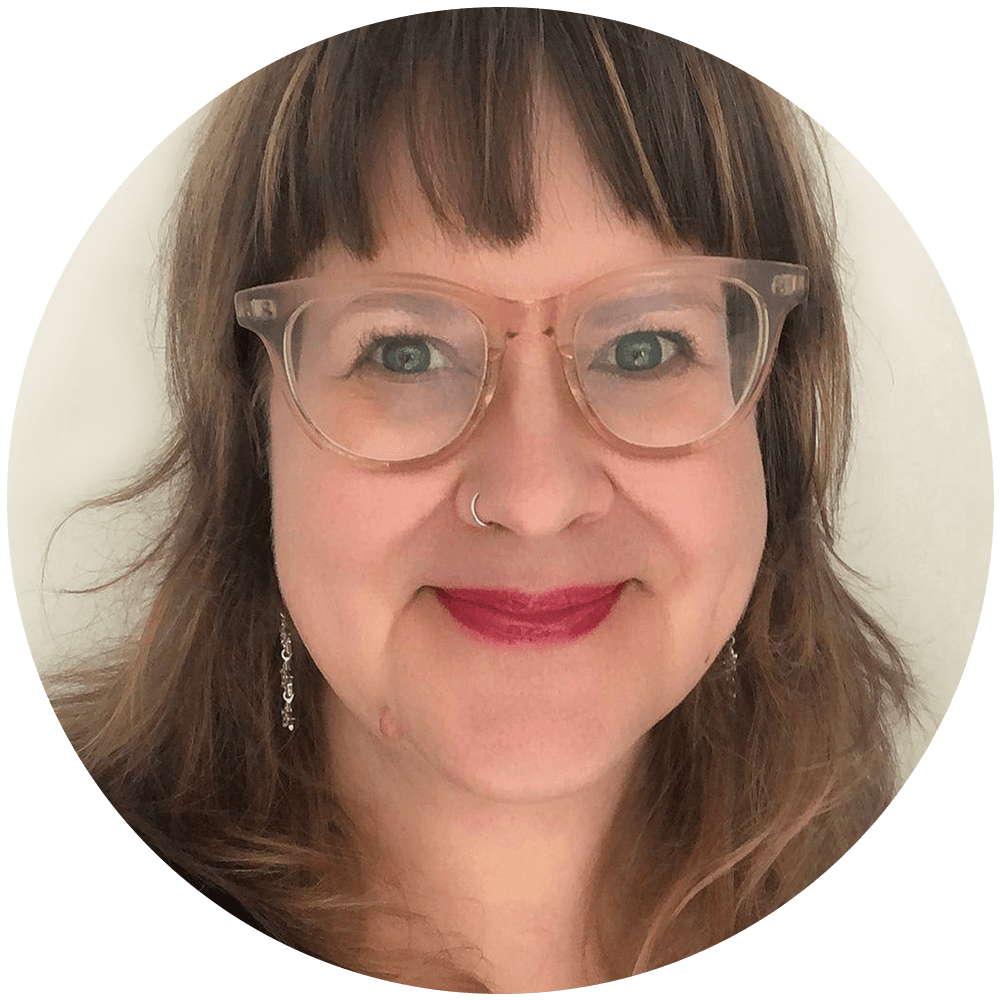 Close up photo of a white woman with medium length brown hair smiling at the camera. She is wearing pink glasses, a black sweater, and star earrings. The background is a white wall.