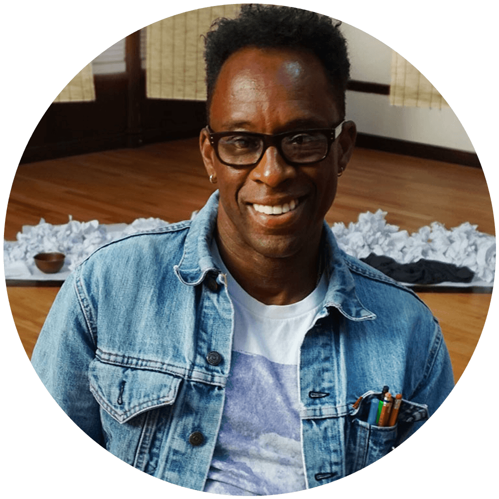 Man with brown skin from the waist up, smiling, wearing thick rimmed black glasses and a denim jacket. He's sitting on an unseen chair in the middle of a large room with a wooden floor. He has a pencil in his hand and pencils in the right pocket of his denim jacket. In a row behind him is a pile of crumpled white typing paper.