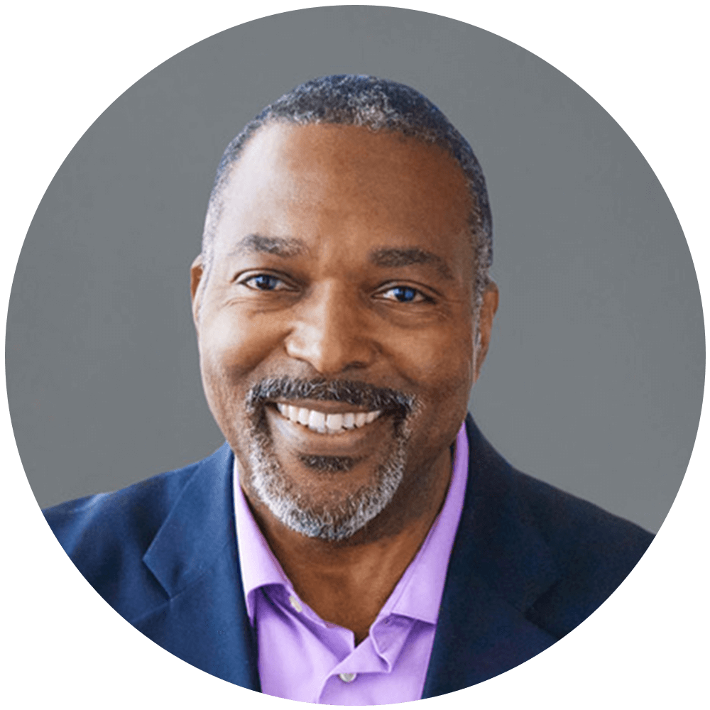 Ted Russell headshot. A black man with salt and pepper short hair and beard smiling at the camera. Wearing a blue blazer and a lilac button up shirt