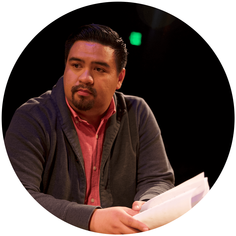 A brown skinned man with black hair in a grey sweater and pink shirt holding a white script at a table.