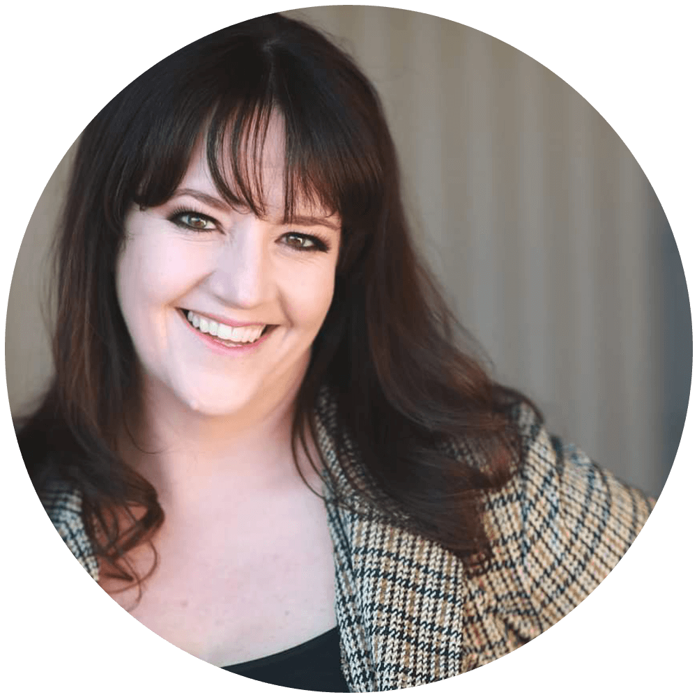 The image shows a Caucasian woman smiling for the camera. She has long layered brown hair with bangs and is wearing and brown and black plaid jacket. She is standing in front of a striped wall.