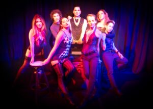 7 actors in Cabaret costumes pose seductively in red and blue lighting