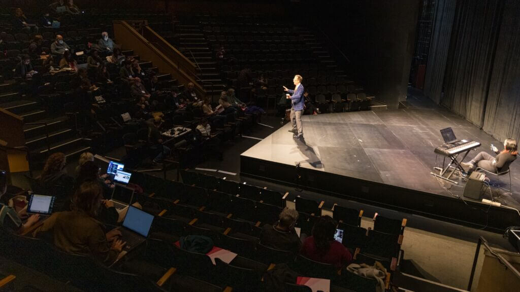 A man stands at the front of the stage with casting directors in the audience.