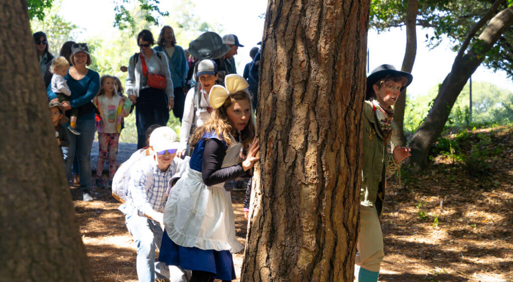 Actors and children from the audience peek out from behind a tree.