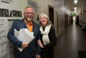 two people with white and grey hair smiling in a backstage hallway