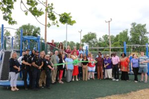A large group stands in front of a newly built accessible playground holding a green ribbon for a ribbon cutting.