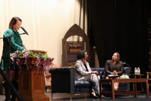 A woman in green stands over a podium posing questions of two women sitting on a couch to her left.