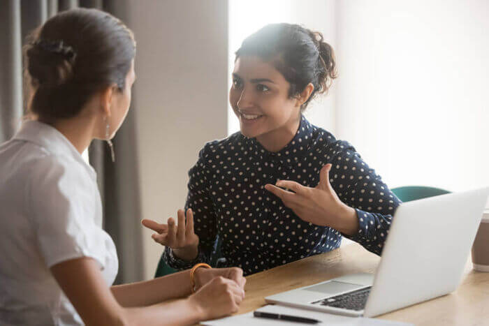 Excited diverse female colleagues discuss ideas at workplace Excited multiethnic female employees discuss work issues sitting at office table, smiling diverse women workers or colleagues engaged in brainstorming talk chatting, explain ideas at workplace